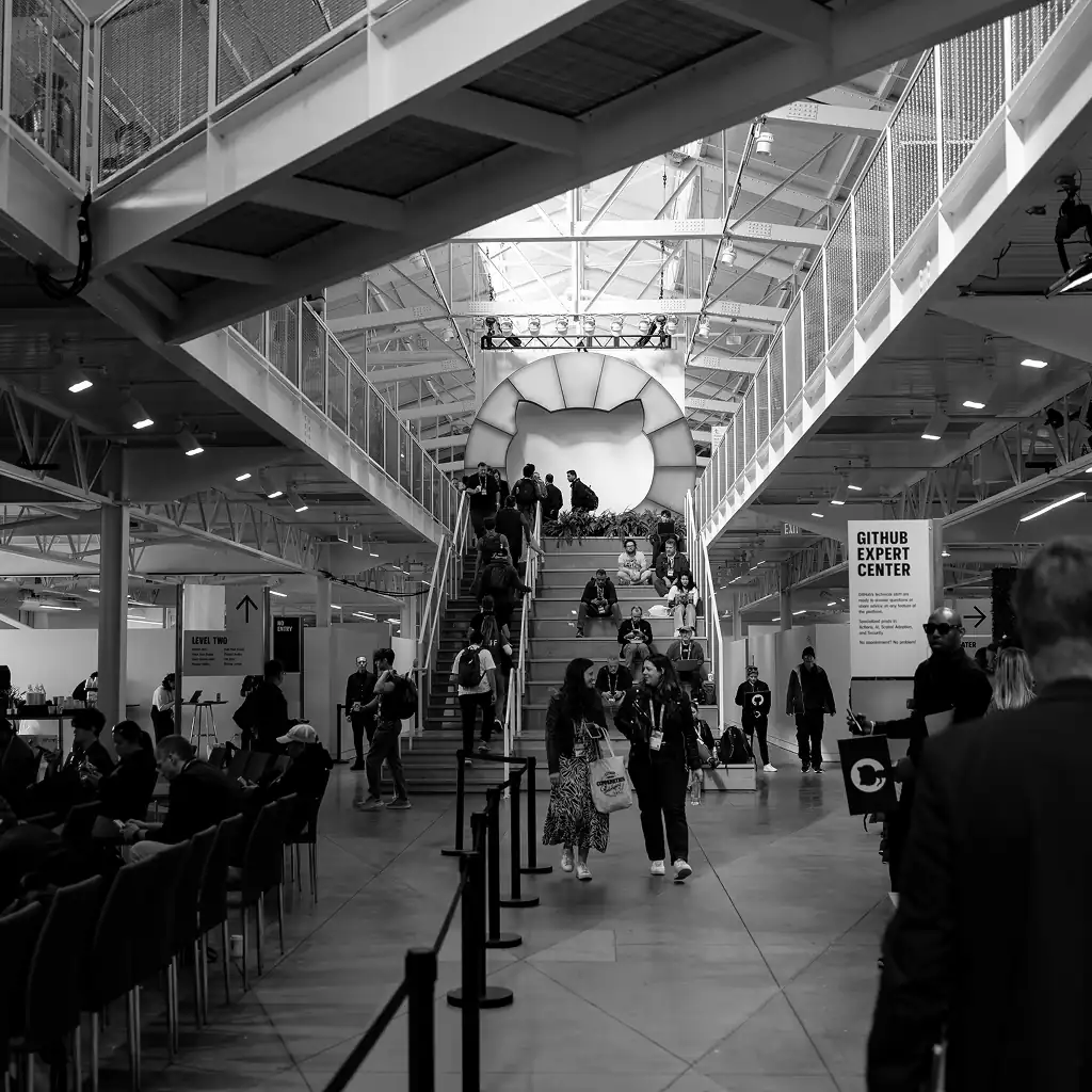 A black-and-white photograph of an indoor event space featuring a staircase with attendees walking and sitting. At the top of the stairs is a large GitHub Octocat logo illuminated in the background. To the right, there is a sign labeled "GitHub Expert Center," inviting attendees to seek assistance or information. The space has a modern industrial design with visible beams and open seating areas.