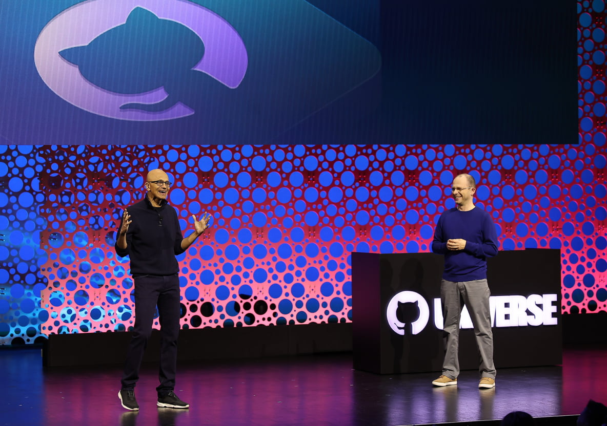 Two speakers on stage at a tech event, one gesturing enthusiastically, with a large display featuring the GitHub logo behind them.