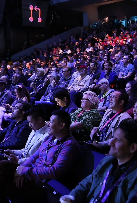 Audience members at a tech conference, attentively watching a presentation, with some participants taking notes.